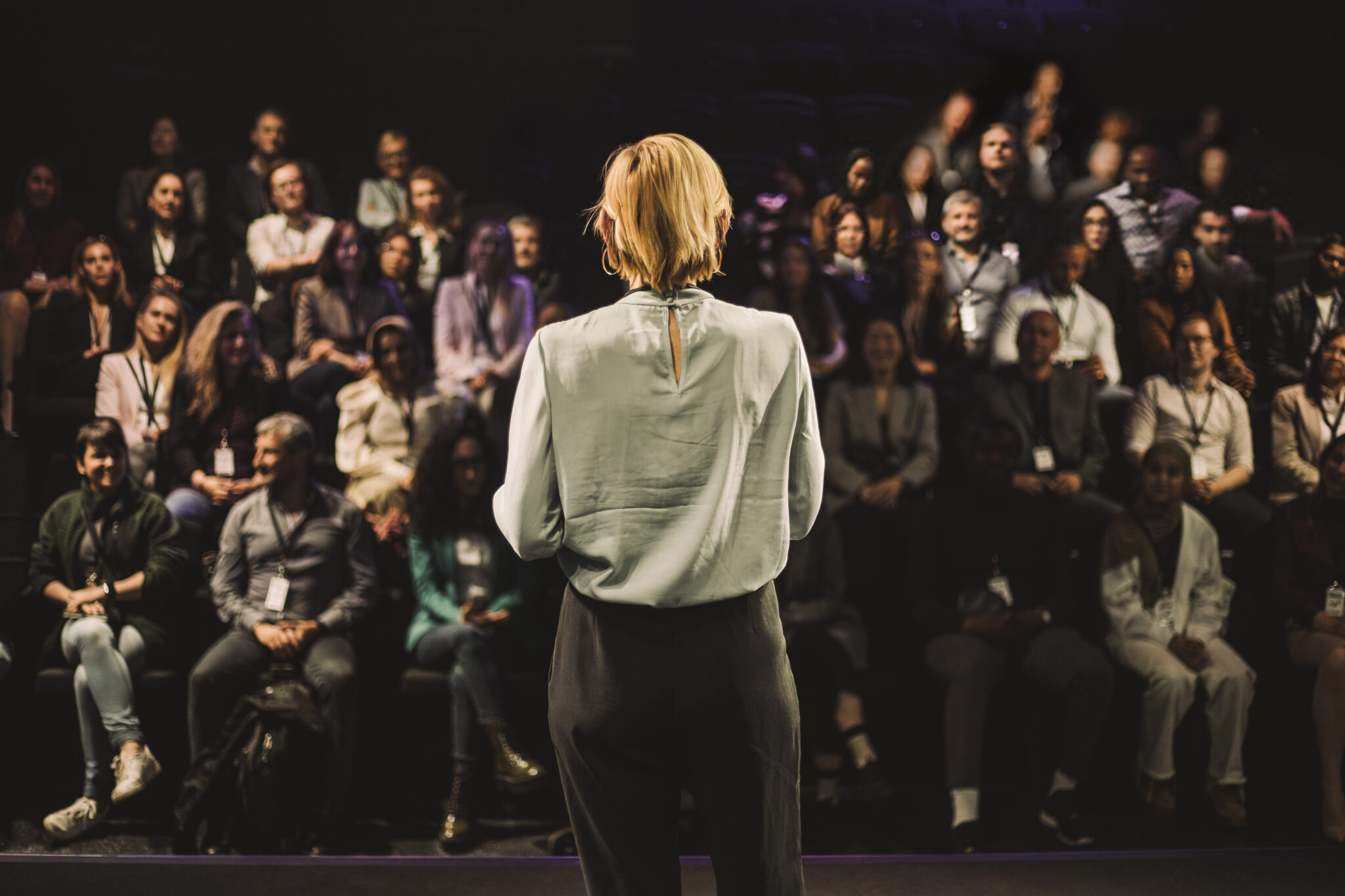 A woman wearing a silk blouse and wool trousers is seen from behind as she stands before an audience in a black box theatre